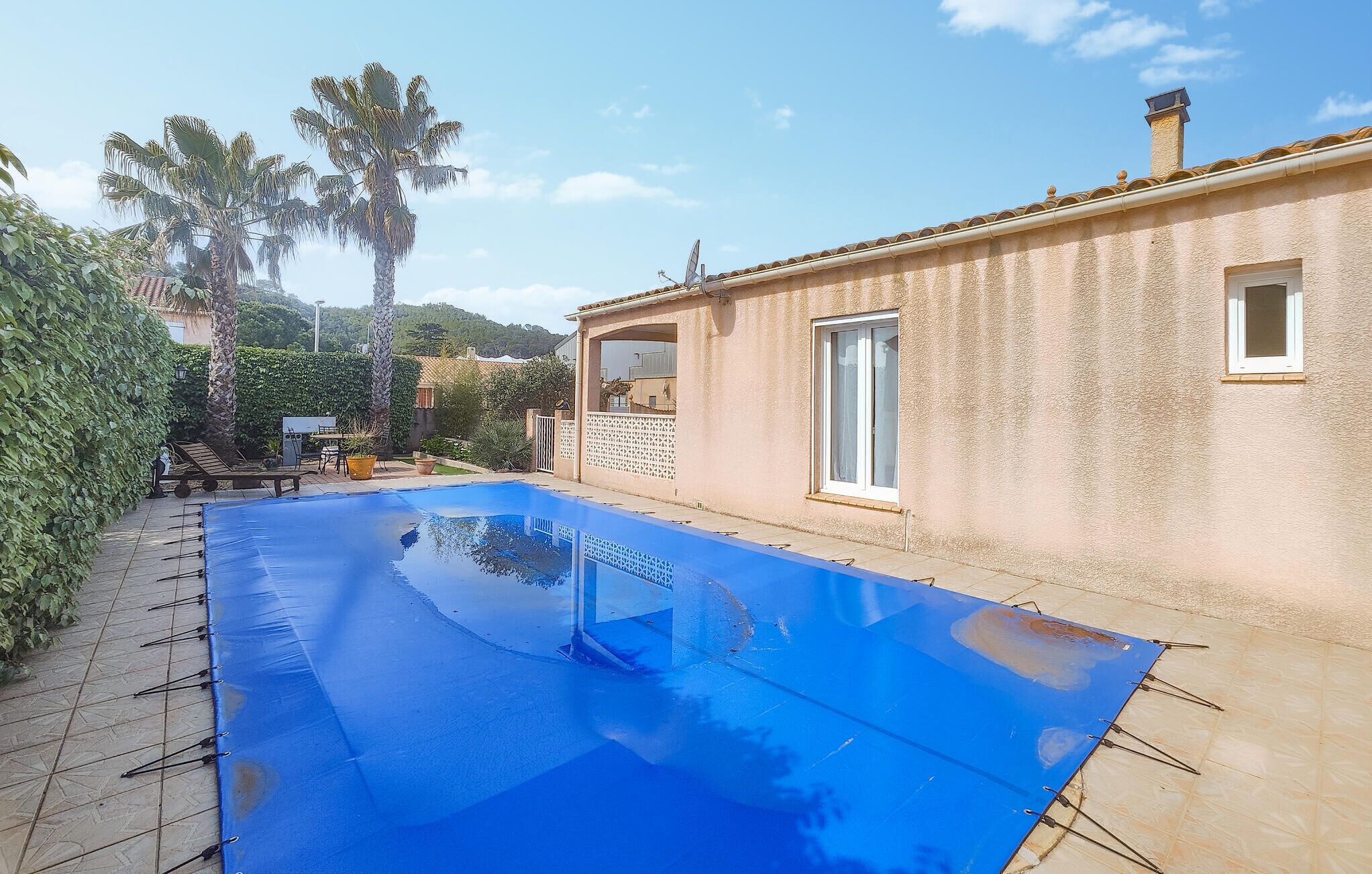 A swimming pool with a blue cover, surrounded by a garden with palm trees and a building with a tiled roof.