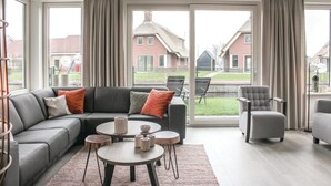 A modern living room with a grey sofa, two round tables, and a grey armchair. Large windows offer a view of a residential area. - 5 bedroom amazing home in Idskenhuizen (Idskenhuizen)