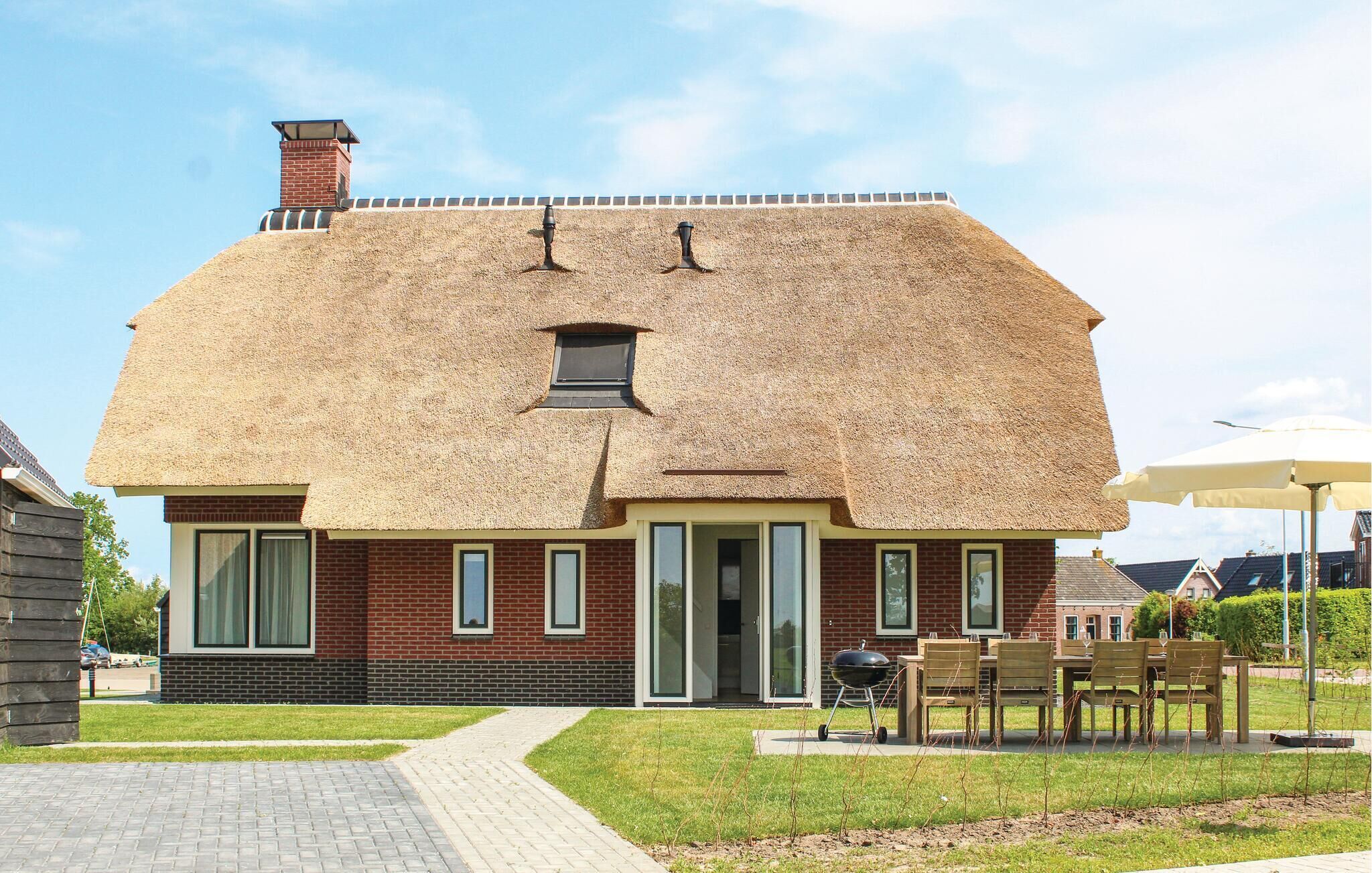 Une maison avec un toit de chaume, une façade en briques et une allée pavée menant à une terrasse en bois aménagée avec du mobilier d’extérieur.