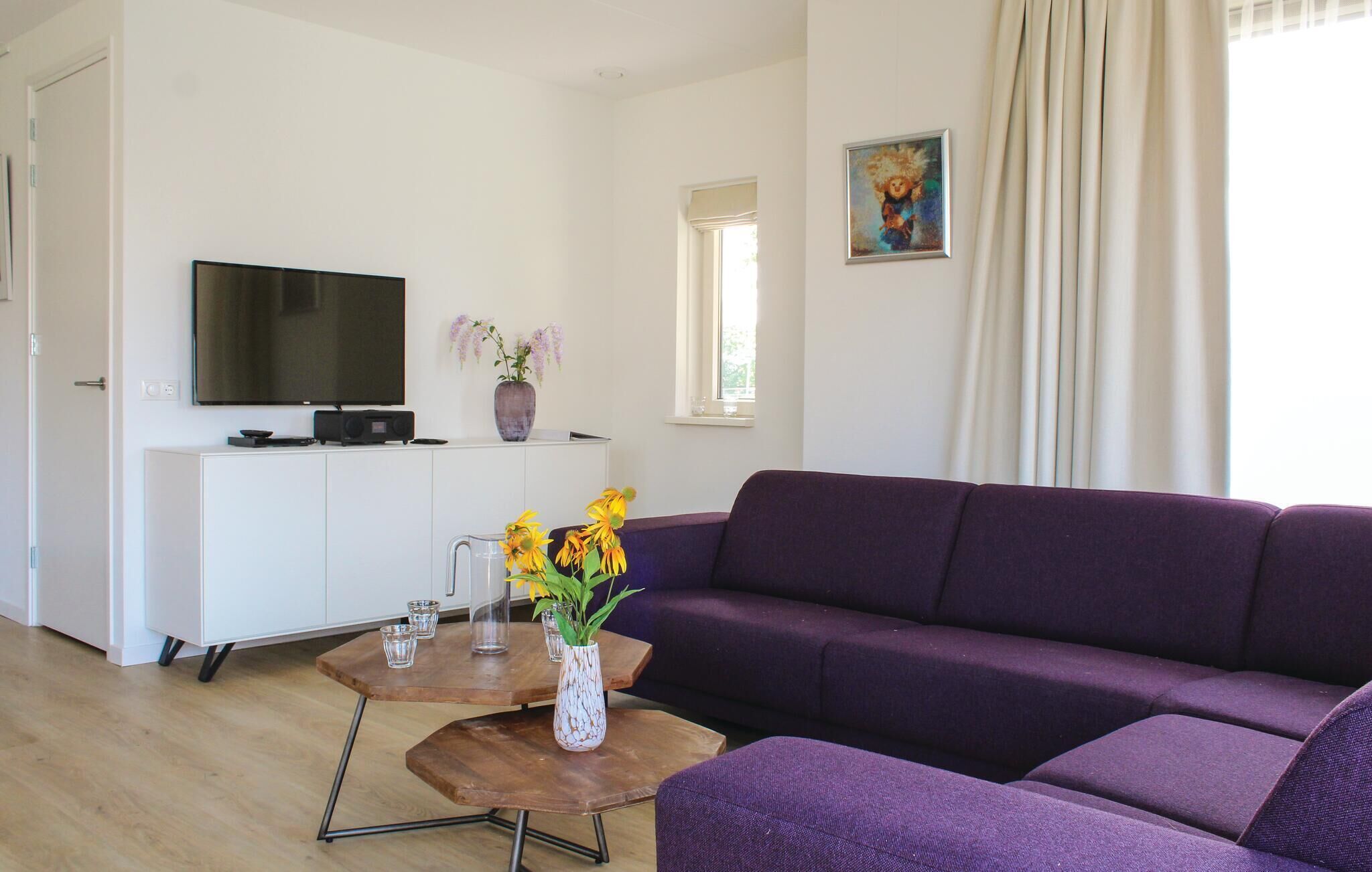A living room with a purple sofa, a coffee table with vases, a flat-screen TV on a white cabinet, and a window with curtains.