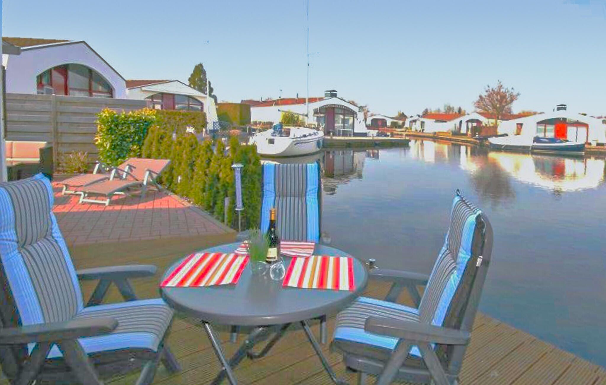 Eine Terrasse mit zwei gestreiften Stühlen und einem Tisch, mit Blick auf ein ruhiges Gewässer und Boote.