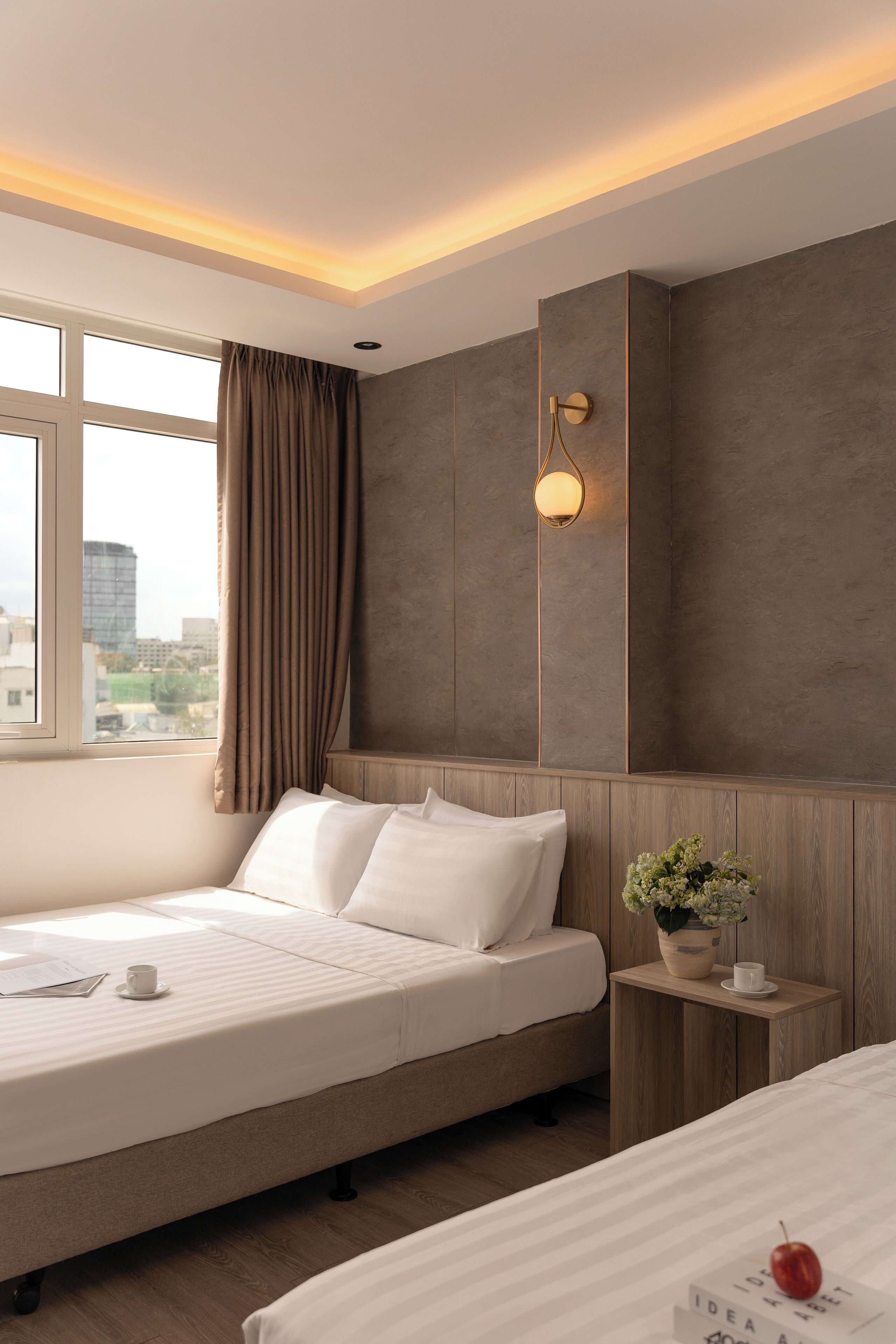 Een moderne hotelkamer met een groot bed, een klein nachtkastje met een plant, een raam met gordijnen en uitzicht op de stad.