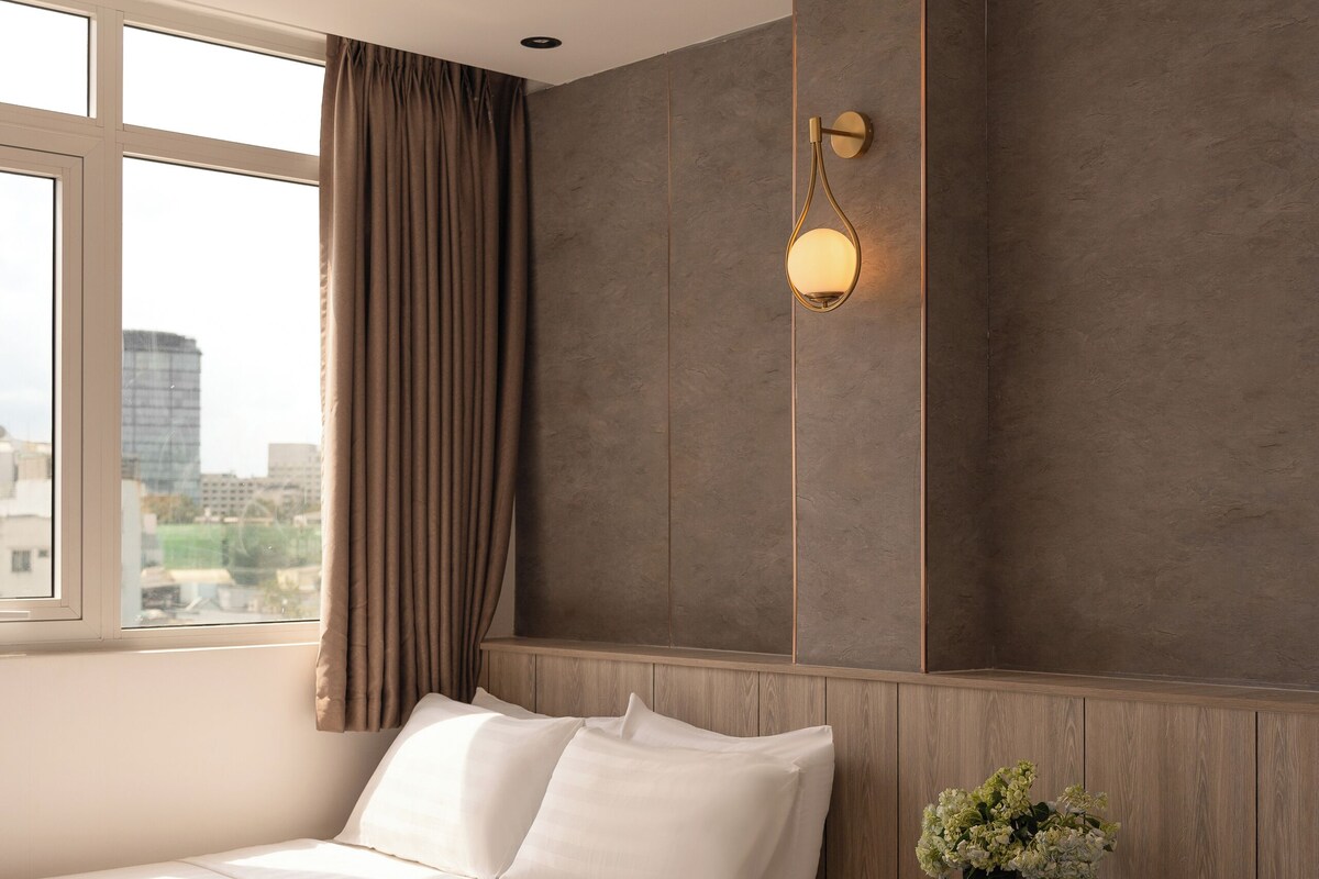 A modern hotel room with a large bed, a small bedside table with a plant, a window with curtains, and a city view.