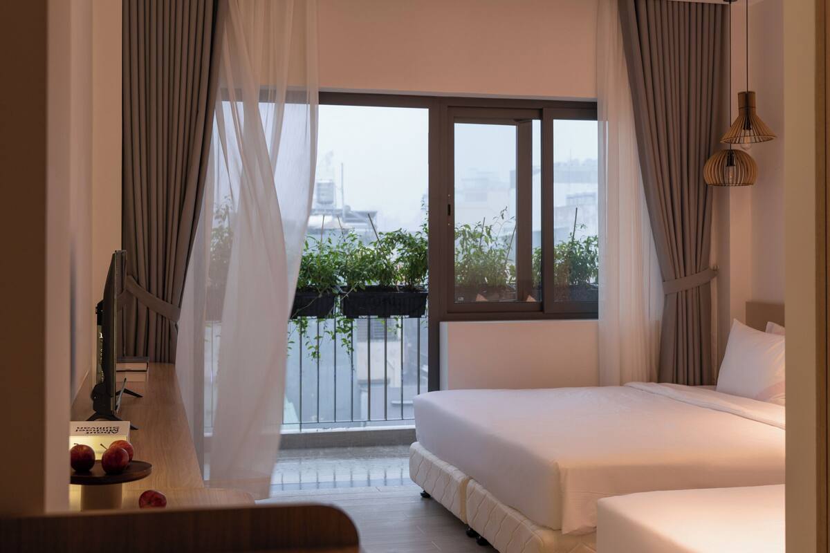A hotel room with a bed, a desk with a cup, and a view of the city through large windows.