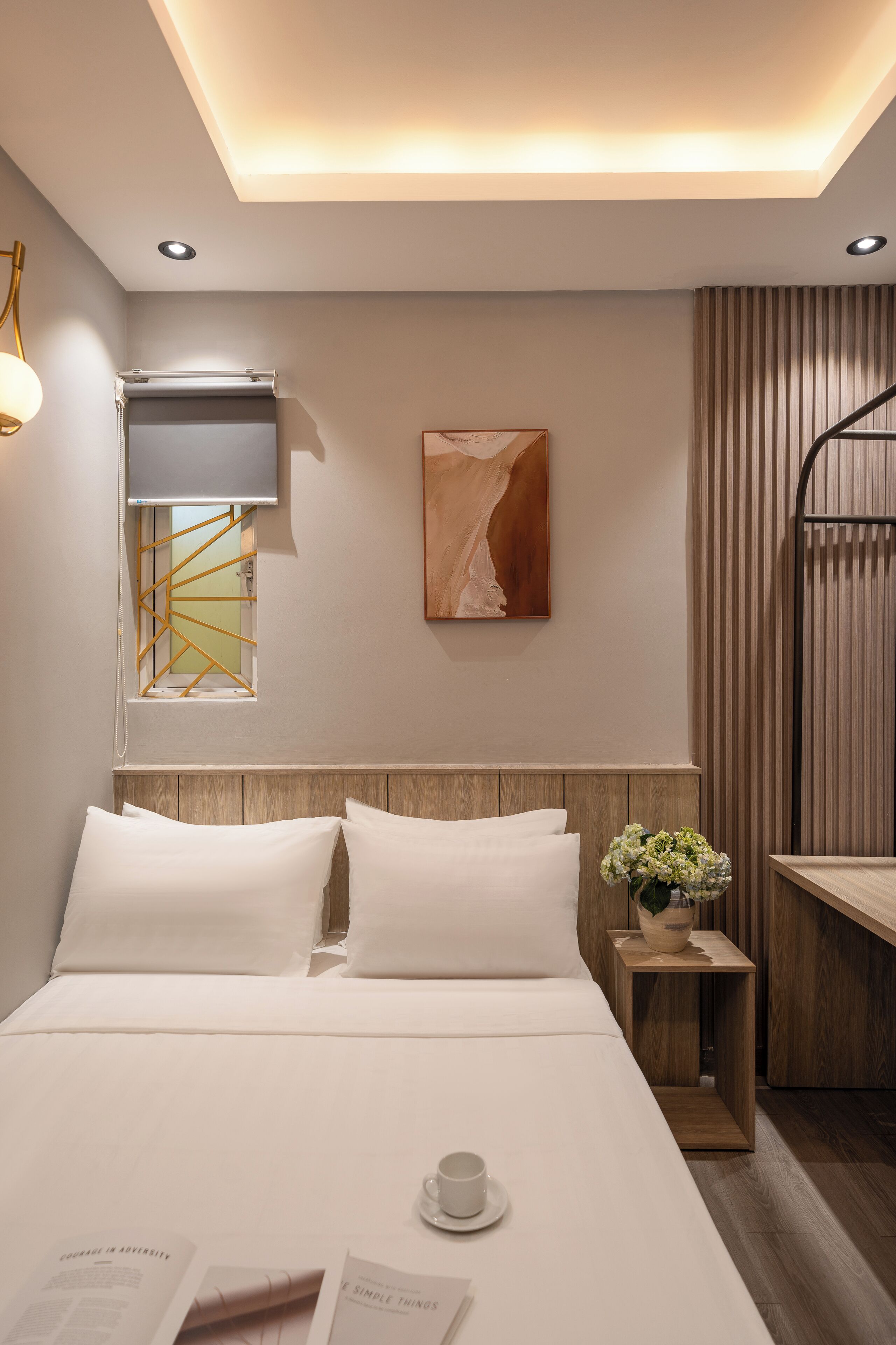 Una habitación de hotel moderna con una cama bien hecha, una mesita de noche pequeña con una taza y un libro, un televisor montado en la pared y una ventana con un diseño geométrico.