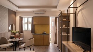 Een kleine hotelkamer met een bed, een eettafel met stoelen, een televisie op een houten standaard, een keuken met kastjes en een groot raam.