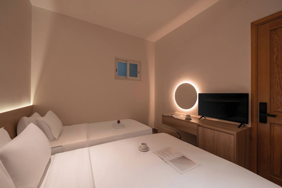 A hotel room with two beds, a TV, a bedside table with a lamp, and a small window.