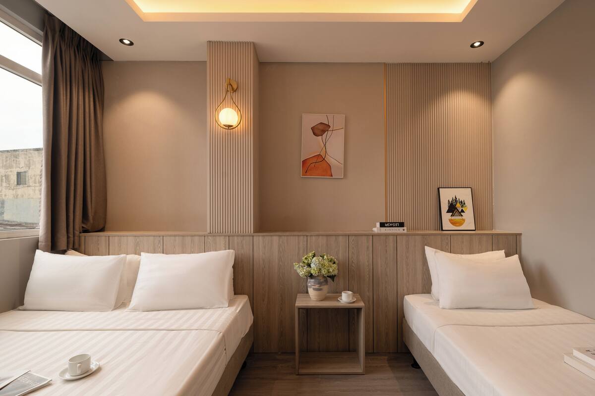 A hotel room with two beds, a small table with a vase of flowers, and a wall-mounted light fixture.