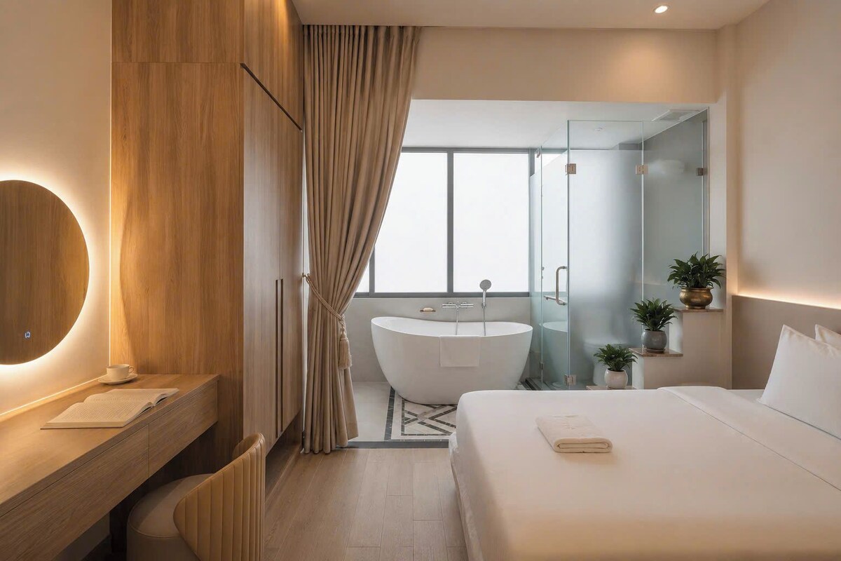 A modern hotel room with a large bed, a round mirror, a vanity with a sink, and a glass-enclosed shower area.