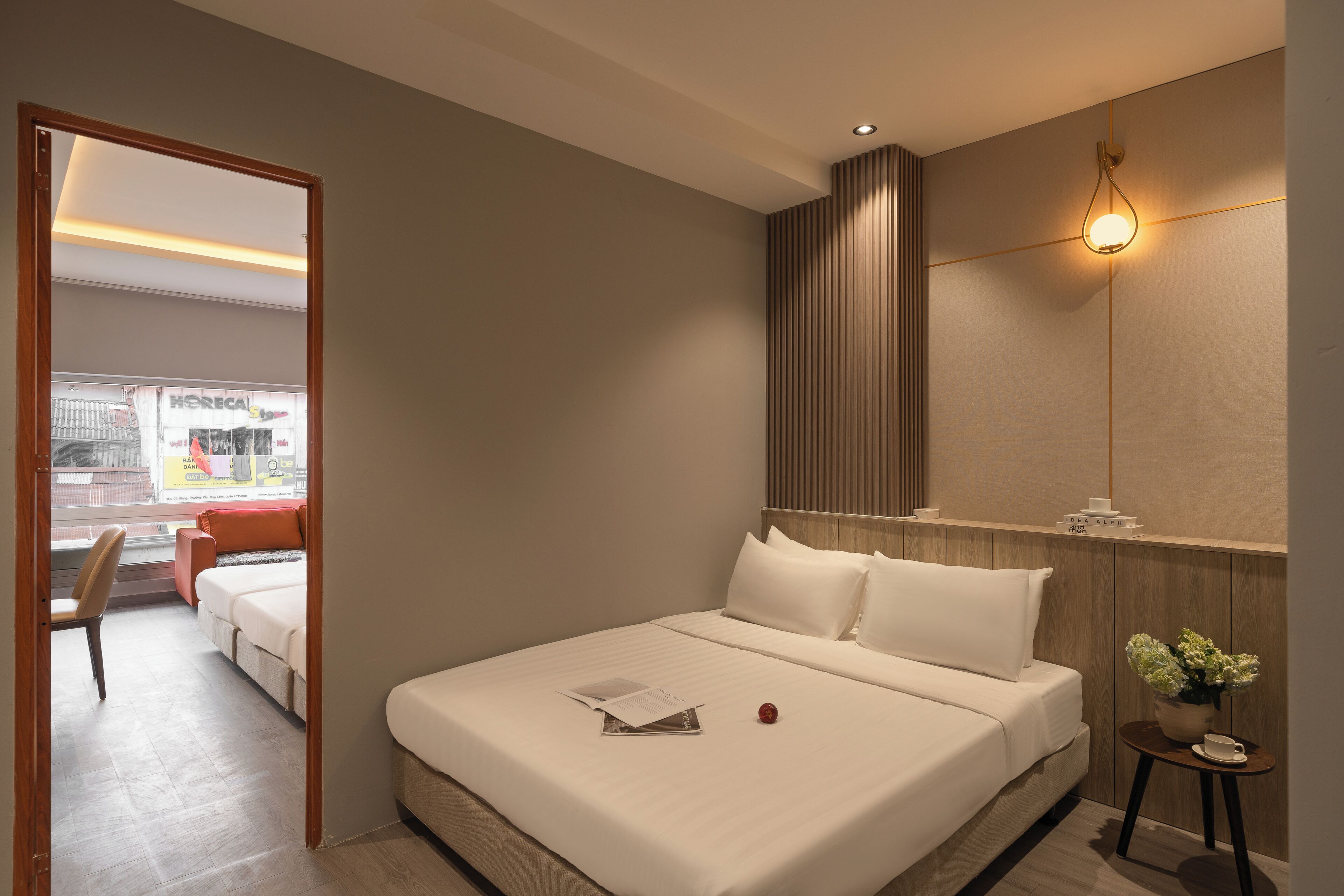 Una moderna camera d'albergo con un letto grande, un comodino con una lampada, un tavolino con una pianta e vista sulla città attraverso una finestra.