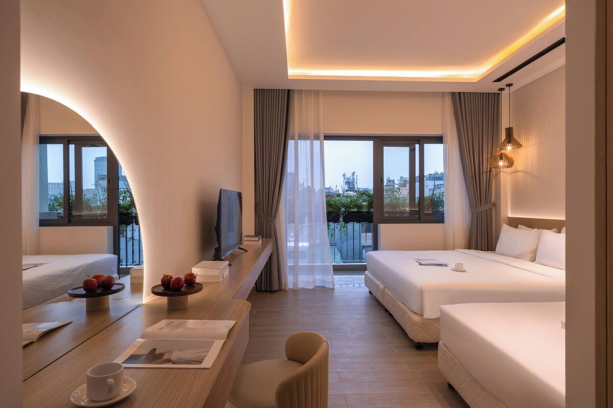 A modern hotel room with two beds, a TV, a dining area with a table and chairs, and large windows offering a city view.
