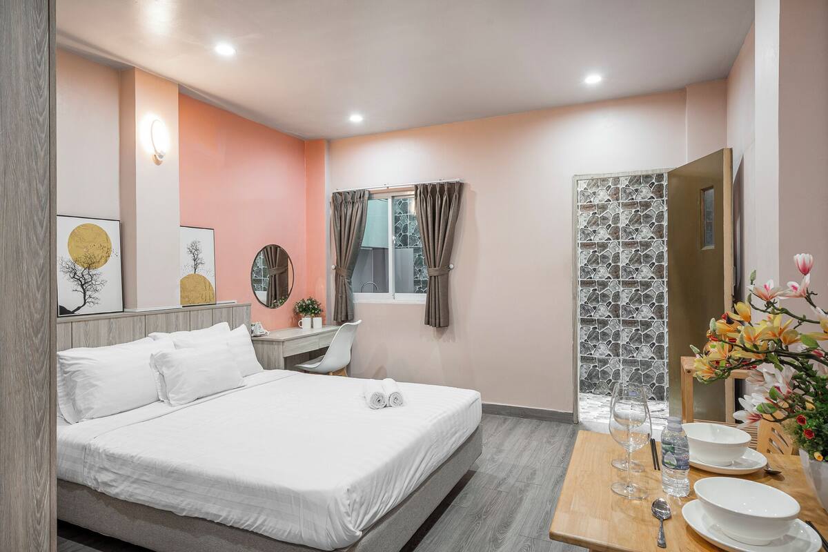 A hotel room with a bed, a desk, a chair, a vanity with a mirror, a window with curtains, a shower area, and a dining table set with plates, glasses, and a vase of flowers.