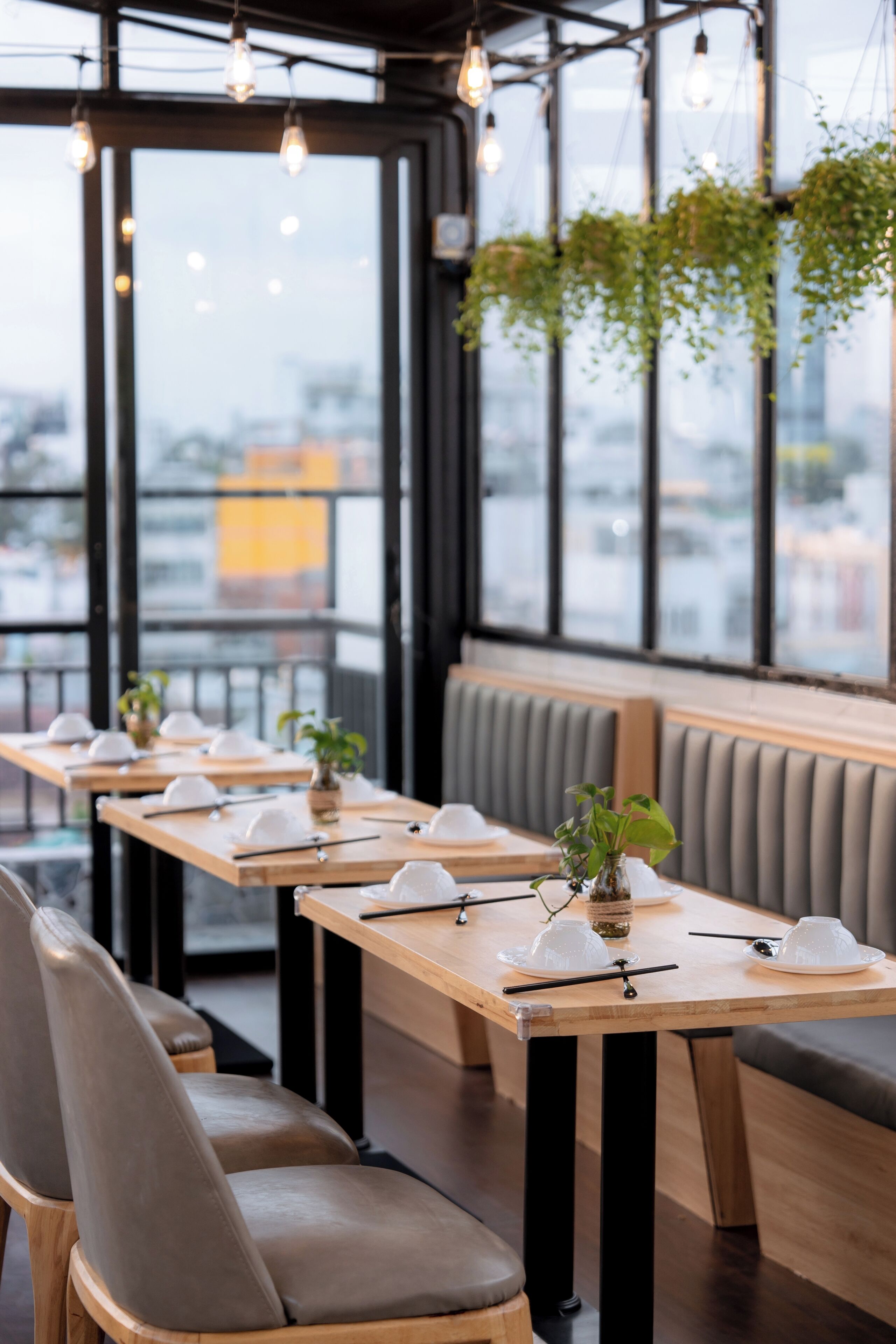 Un restaurant avec de longues tables en bois, des sièges rembourrés de couleur grise et des plantes suspendues.