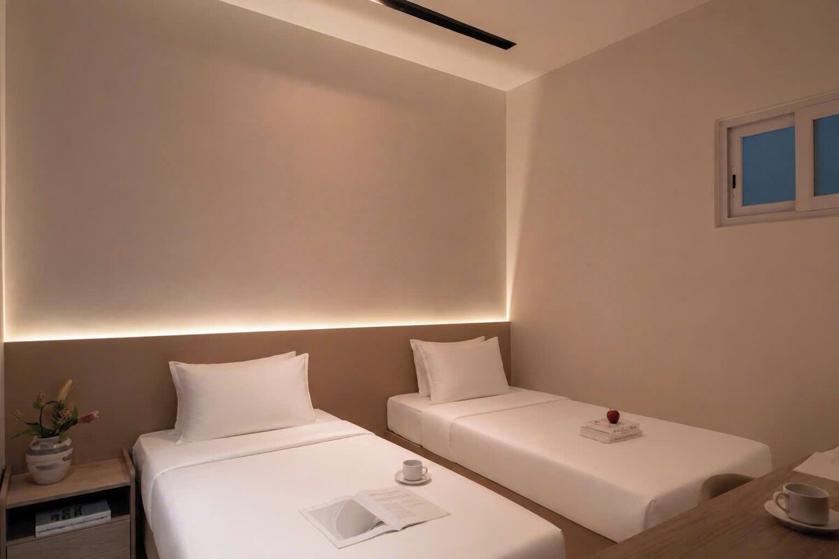 A hotel room with two single beds, a bedside table with a book and a cup, and a window with blinds.