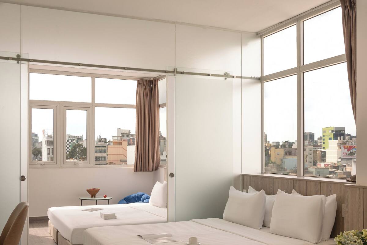 A hotel room with two beds, a desk, a chair, and large windows offering a city view.