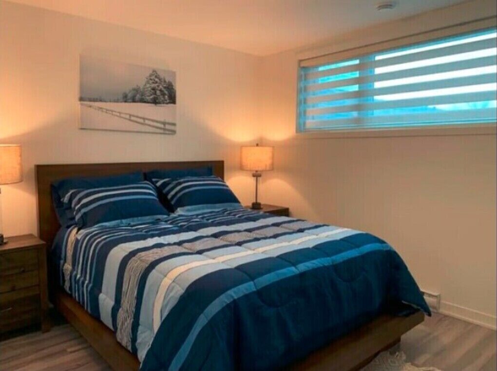 A bedroom with a bed, two bedside tables, a window with blinds, and a wall-mounted picture.