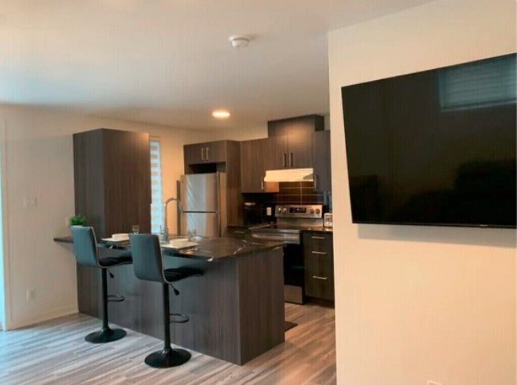 A modern kitchen with dark cabinetry, a central island with bar stools, and a large flat-screen TV mounted on the wall.