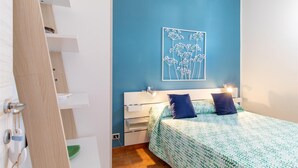 A bedroom with a bed, a nightstand, a wall-mounted lamp, and a blue wall with a white floral artwork. - Beautiful apartment in Marina di Ragusa with house sea view (Marina di Ragusa)
