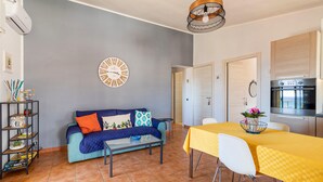 A modern living room with a sofa, a dining table with chairs, a kitchen area with an oven, and a wall clock. - Beautiful apartment in Marina di Ragusa with house sea view (Marina di Ragusa)