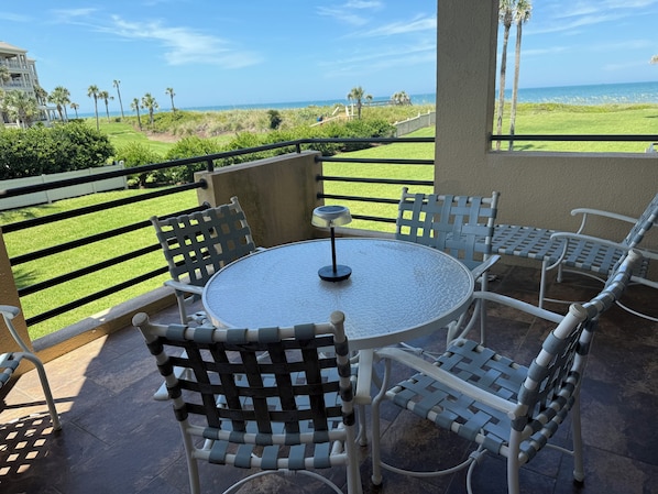A balcony with a round table and four chairs, overlooking a grassy area with palm trees and a view of the ocean.