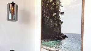 Une chambre à coucher avec un lit, un luminaire suspendu fixé au mur, un tableau représentant un paysage côtier encadré et une plante en pot.