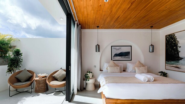Une chambre moderne avec un plafond en bois, un grand lit, deux fauteuils en osier, une petite table d’appoint et une vue sur le ciel par une grande fenêtre.