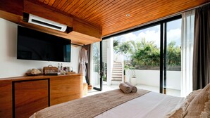 Une chambre avec un plafond en bois, un grand lit, un téléviseur à écran plat fixé au mur et un balcon donnant sur un paysage verdoyant.