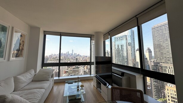 Ein modernes Wohnzimmer mit einem Glastisch, einer Couch, einem Fernseher und Blick auf eine Stadtlandschaft durch große Fenster.
