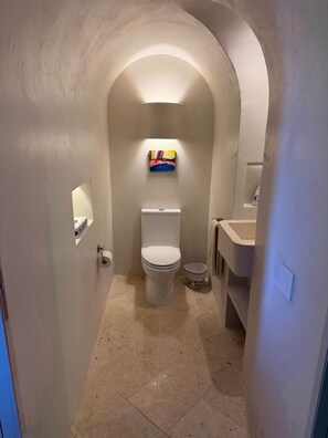 A narrow bathroom with a toilet, a sink, and a small shelf with a colorful item.
