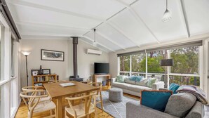 A spacious living area with a wooden dining table, comfortable seating, a sofa, and a large window offering a view of trees. - High Road Haven (Halls Gap)