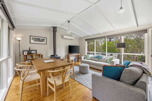 A spacious living area with a wooden dining table, comfortable seating, a sofa, and a large window offering a view of trees.