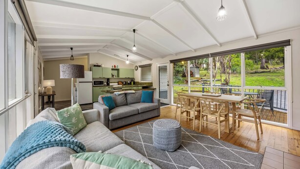 A spacious living area with a sofa, ottoman, and dining table. A kitchen is visible in the background, and a large window offers a view of the outdoors. - High Road Haven (Halls Gap)