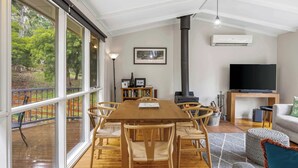 A spacious living area with a large wooden table, comfortable seating, a fireplace, a television, and a view of the outdoors. - High Road Haven (Halls Gap)