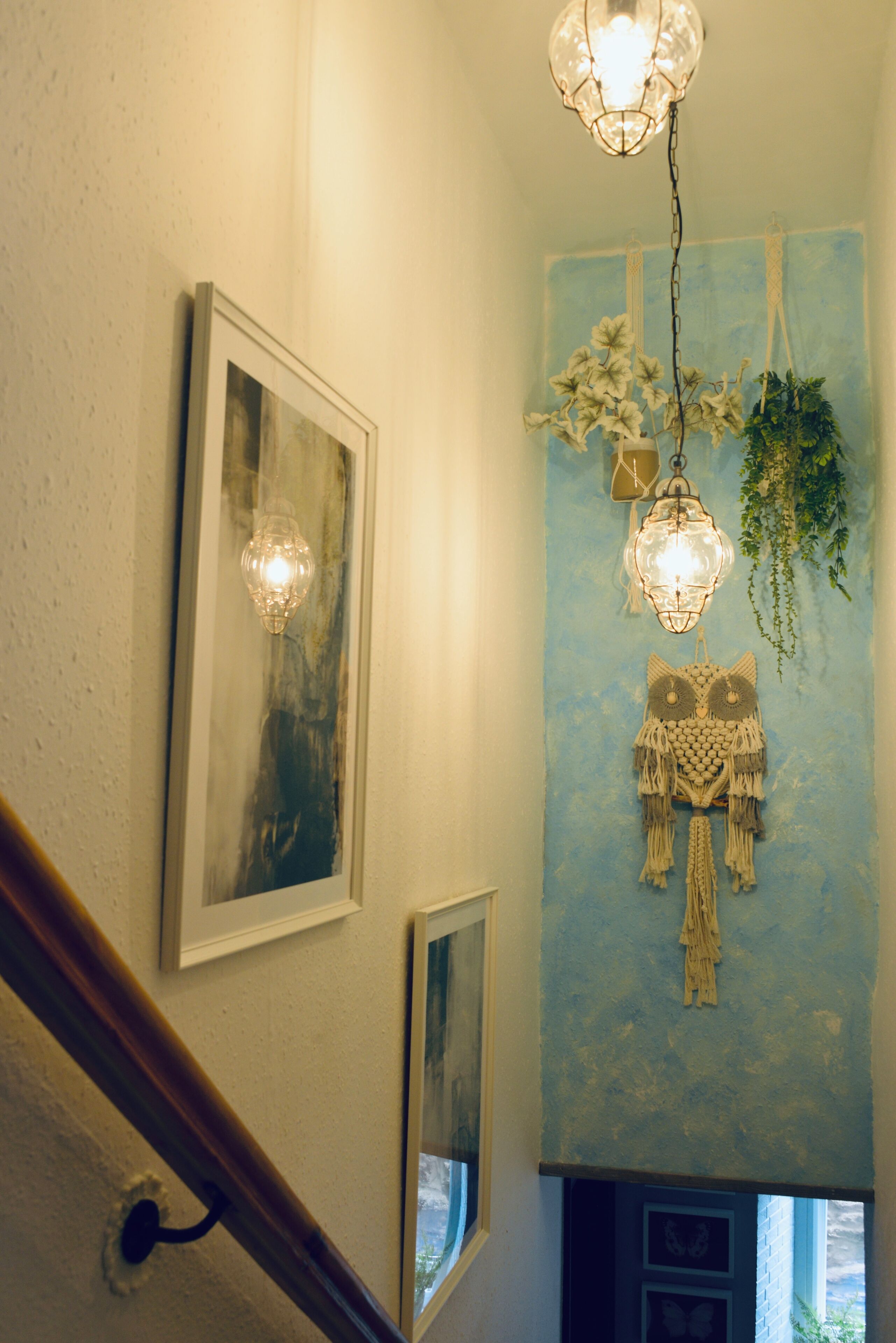 Een trap met houten leuning, een muur met ingelijste foto’s en een decoratieve wandhanger met hangende planten.