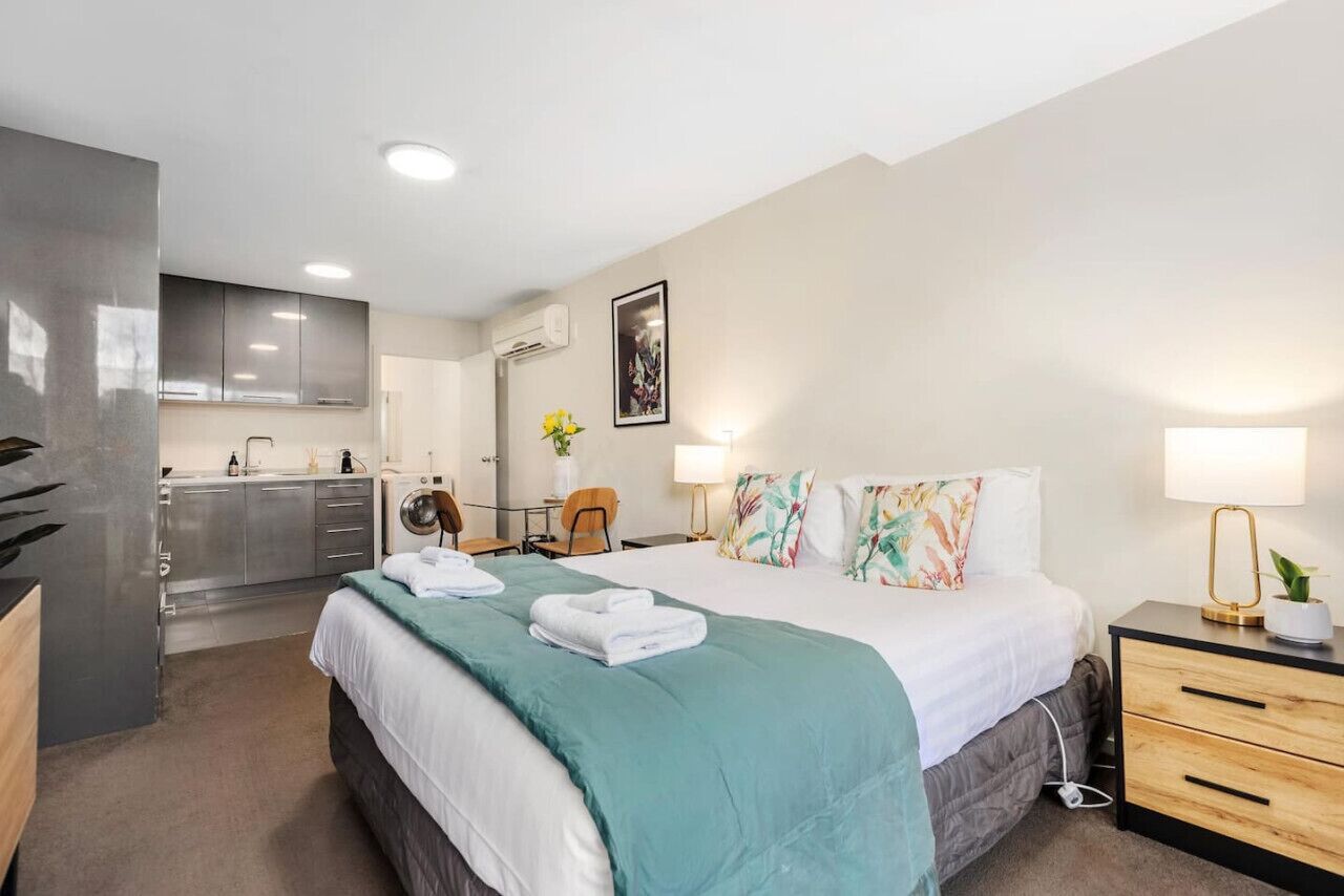 A modern hotel room with a bed, bedside tables, a desk, and a kitchenette.