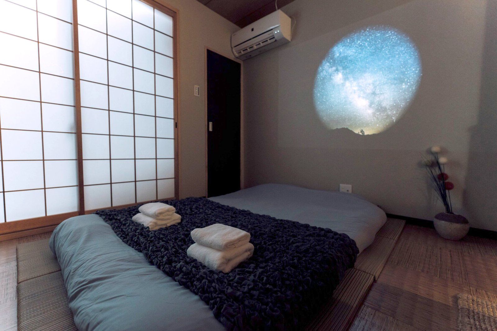 Een kamer met een bed, een futon, een schuifdeur van shoji-papier, een beamer en een kleine plant.