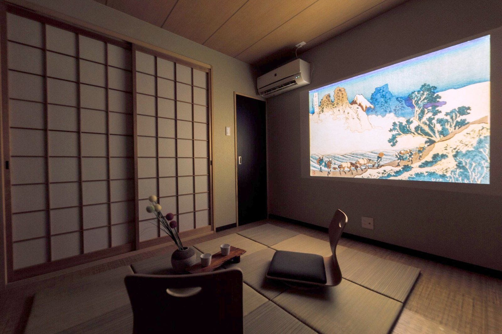 A traditional Japanese room with tatami flooring, a sliding door, a projector screen displaying a landscape, and a small table with a vase and cups.