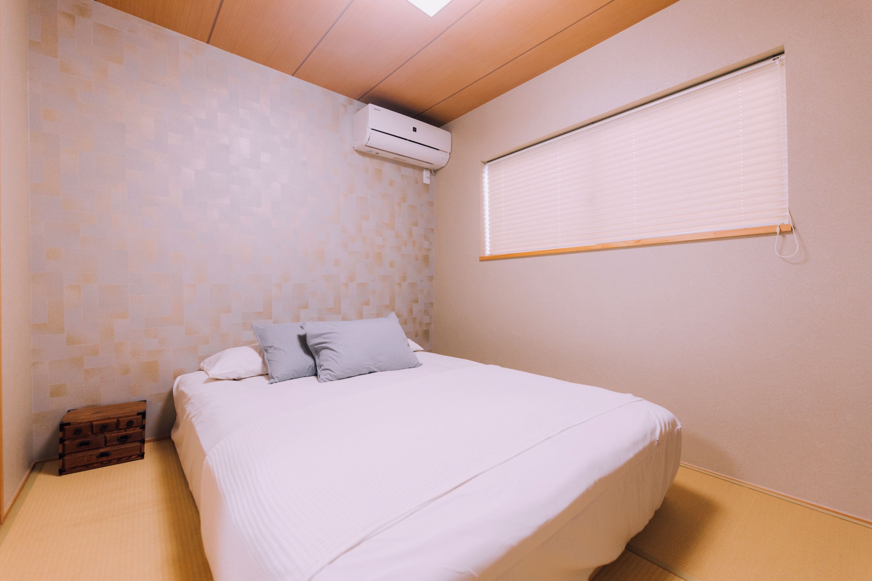 A bedroom with a bed, pillows, a wooden chest, an air conditioning unit, and a window with blinds.