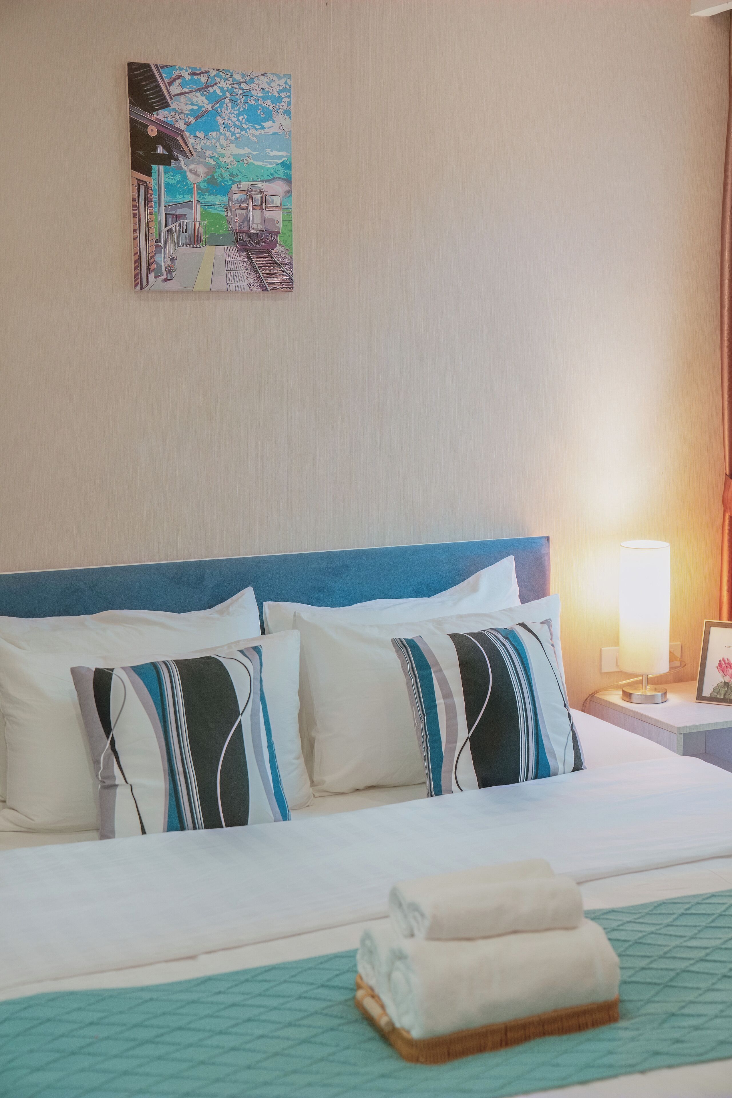 A hotel room with a bed, pillows, a bedside table with a lamp, and a painting on the wall.