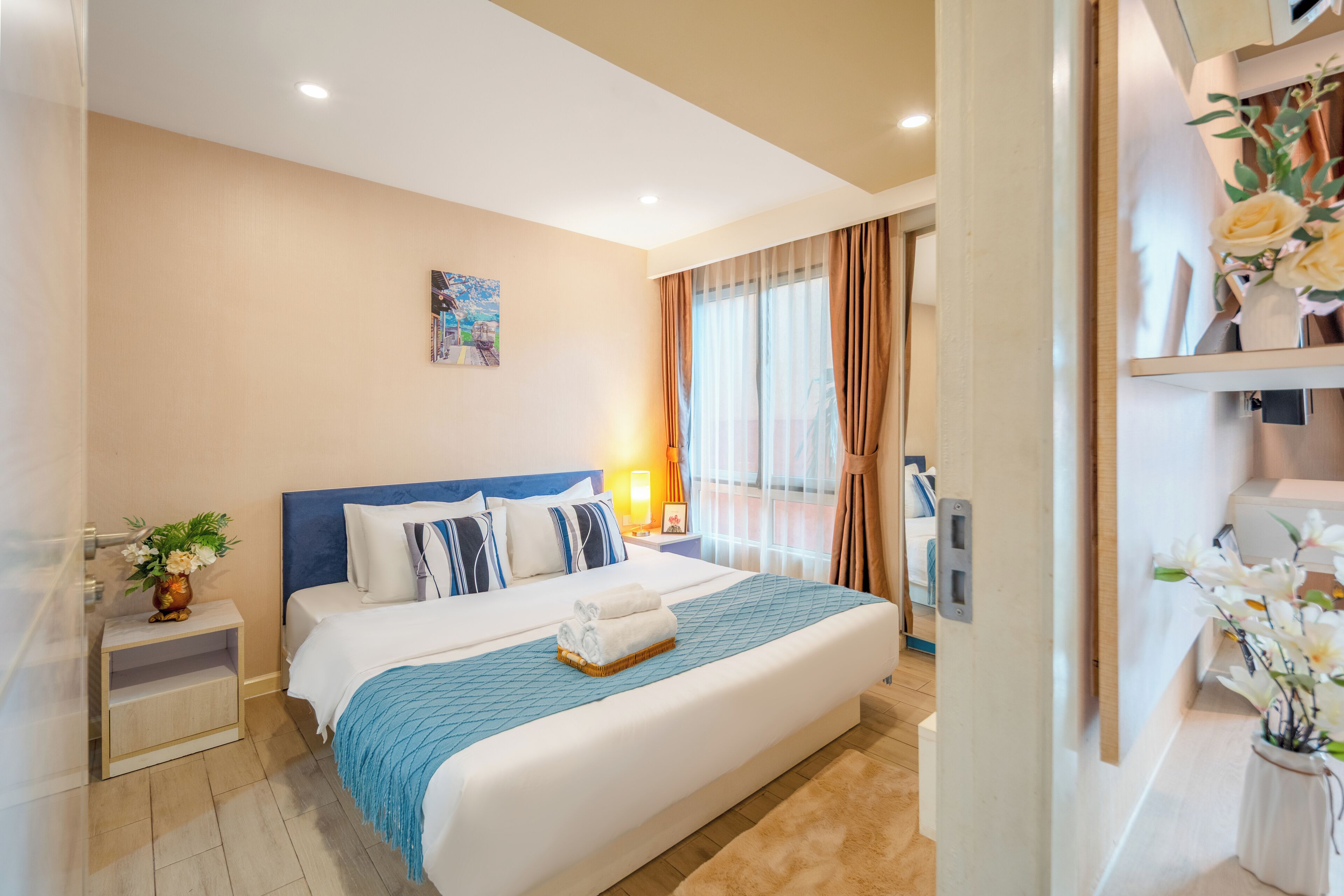 A hotel room with a large bed, a bedside table with a vase, a nightstand, a window with curtains, and a wall-mounted TV.