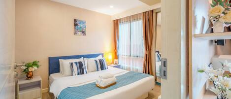 A hotel room with a large bed, a bedside table with a vase, a nightstand, a window with curtains, and a wall-mounted TV.