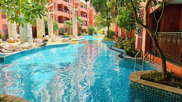 A swimming pool with clear blue water, surrounded by tropical plants and a red building with balconies. - Seven Seas Condo Resort steps to  Beach (Pattaya)