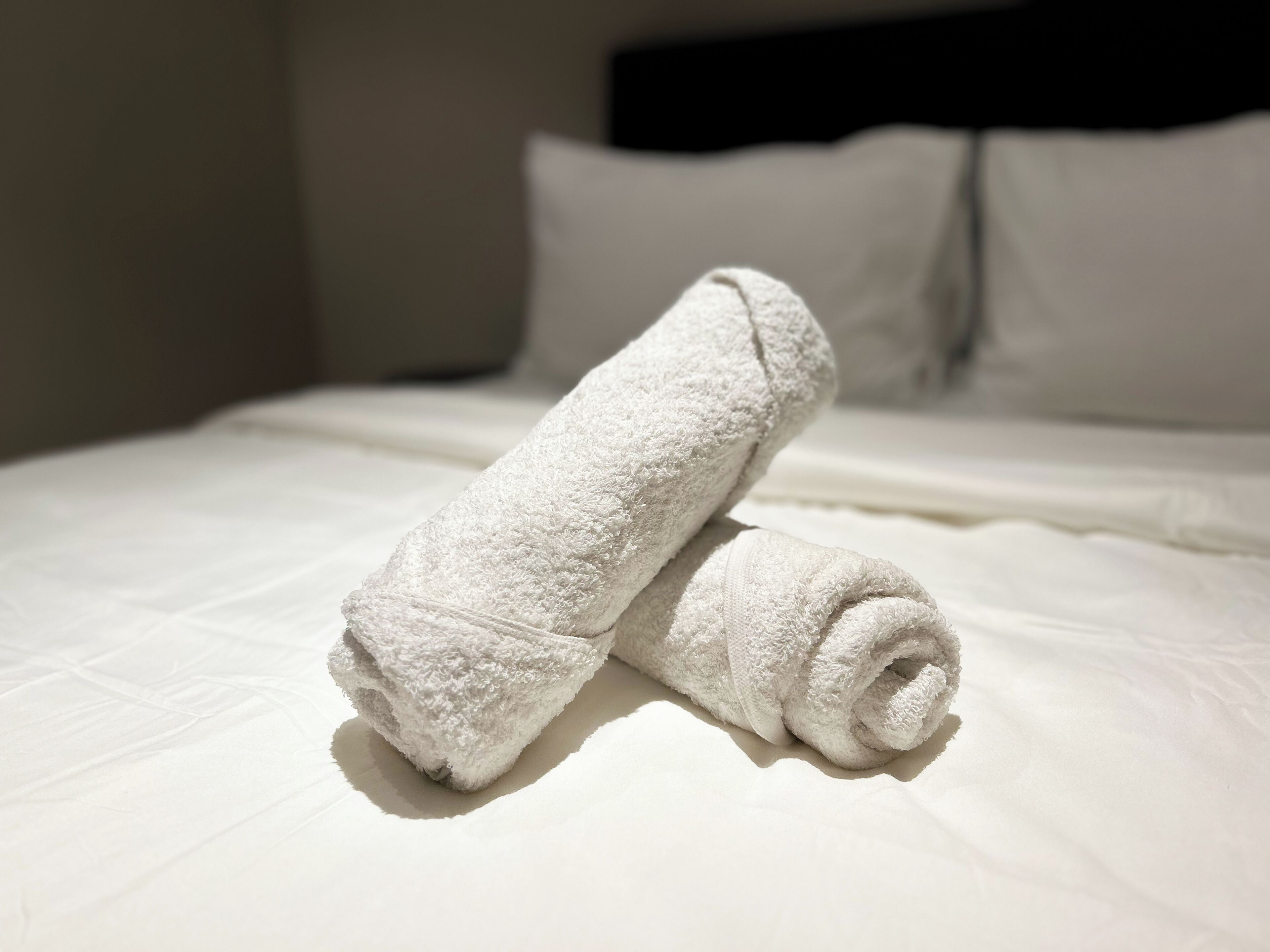 A neatly made bed with white linens and two rolled towels placed on it.