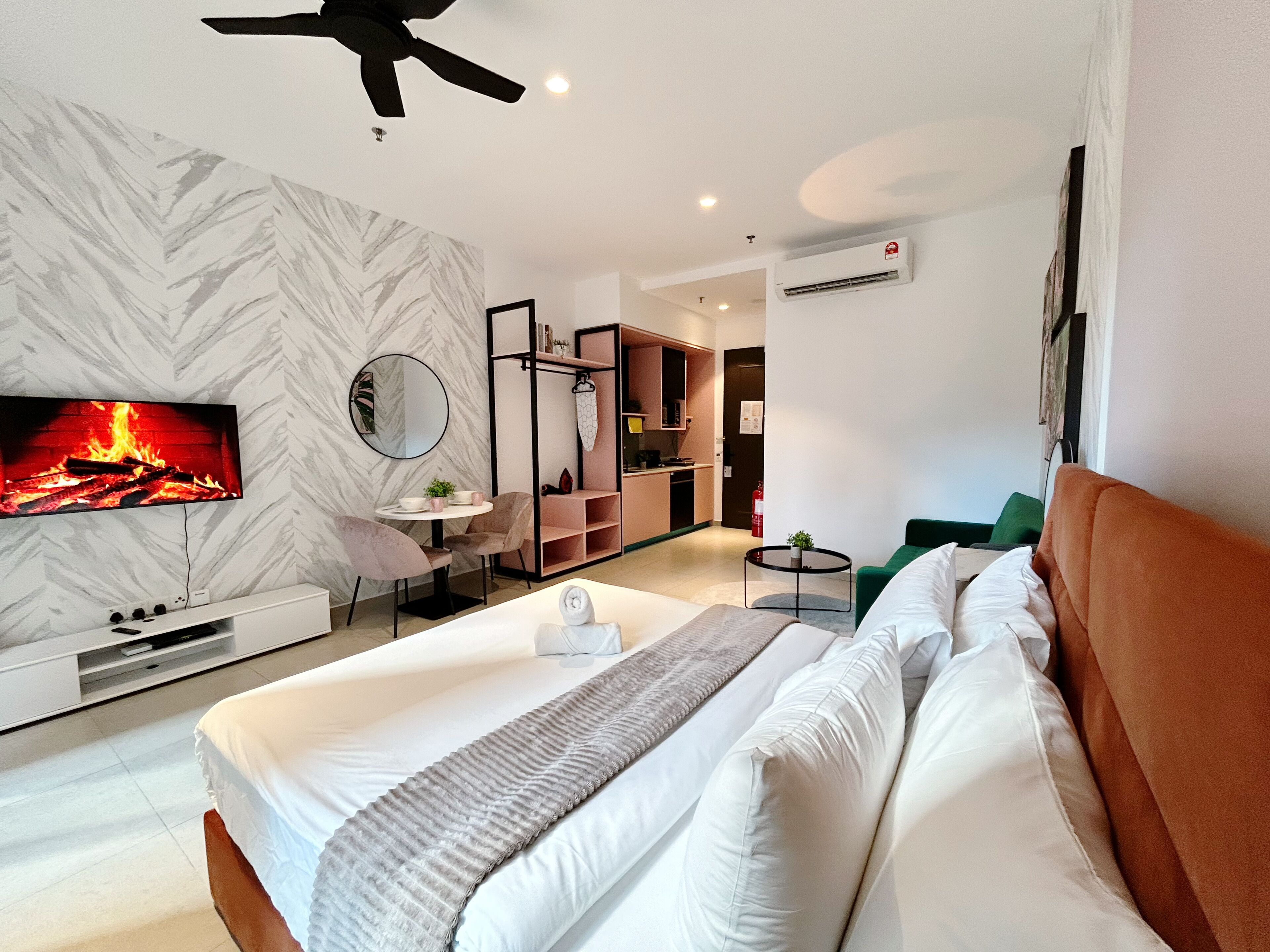 A modern hotel room with a large bed, a television displaying a fireplace, a kitchenette, and a ceiling fan.