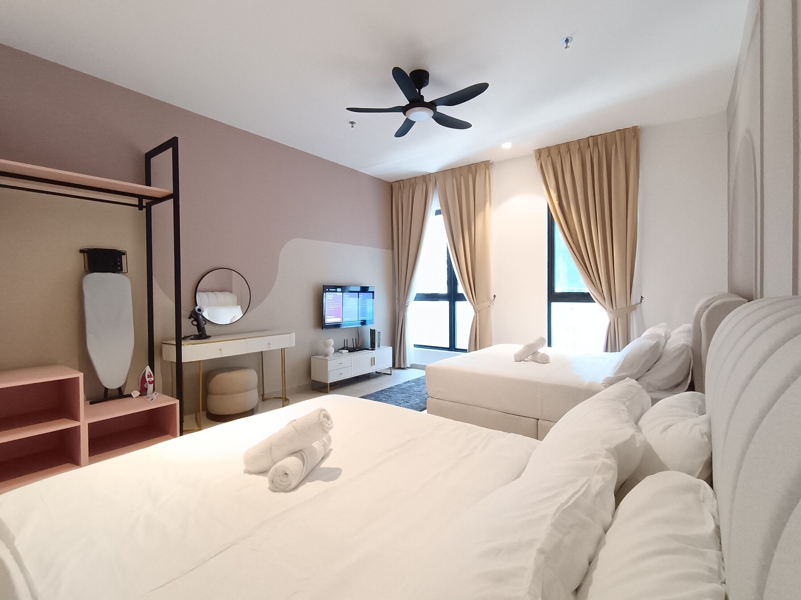 Une chambre d’hôtel avec deux lits, un bureau avec une télévision, un ventilateur de plafond et de grandes fenêtres avec des rideaux.