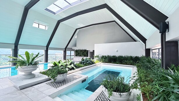 Un área de piscina interior moderna con diseño geométrico en el techo, plantas en macetas y una vista despejada del paisaje circundante.