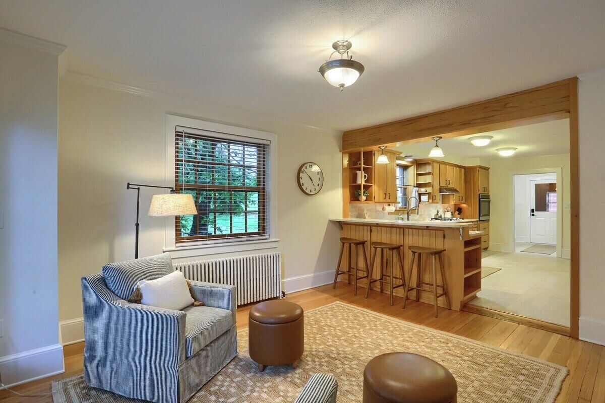 A living room with a sofa, ottoman, and a lamp near a window. There is a kitchen area with bar stools and a refrigerator.