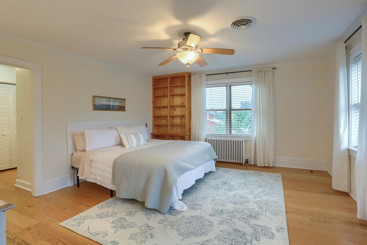 Une chambre à coucher avec un lit, un ventilateur de plafond, une fenêtre avec des rideaux et un radiateur.