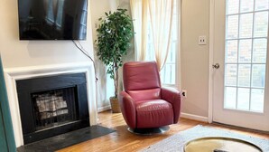 Una sala de estar con chimenea, un sillón rojo, una mesa de centro redonda y un ventanal con cortinas.