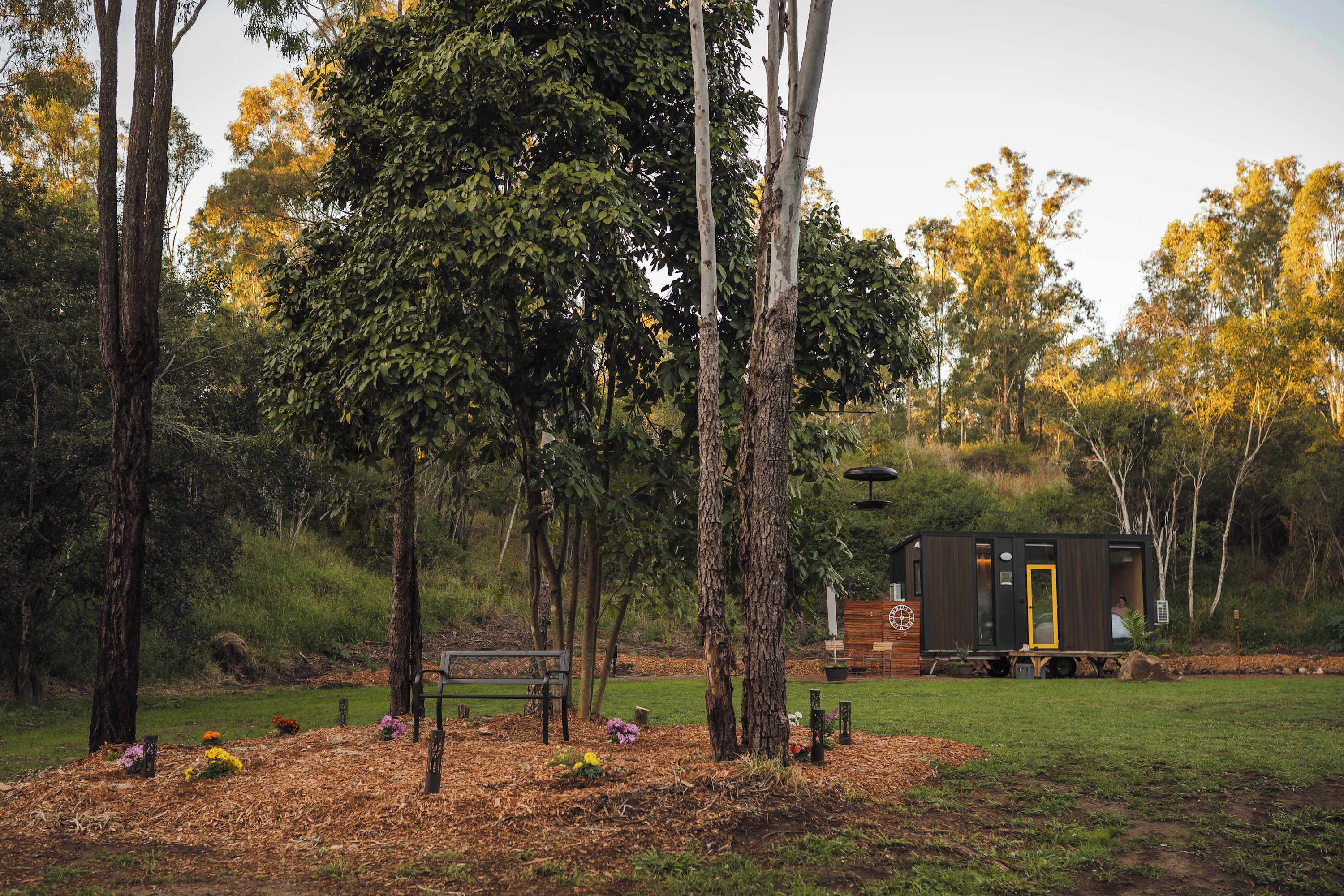 A rustic cabin with a wooden deck and a bench, surrounded by trees and a grassy area.